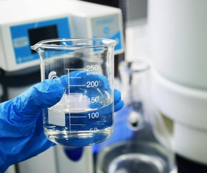 Gloved hand holding a beaker with clear liquid, set against a laboratory background.